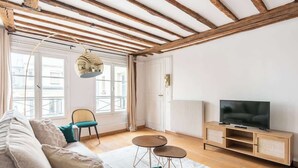 Uma sala de estar com um sofá, duas poltronas, uma televisão sobre um móvel de madeira e uma janela grande com cortinas.