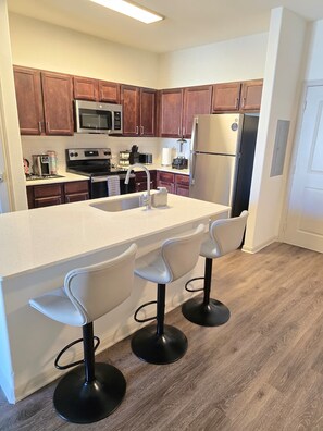 A modern kitchen with wooden cabinets, stainless steel appliances, a central island with bar stools, and a wooden floor. - LUXURY 1-bedroom apartment in superb Frisco near Toyota Stadium. (Frisco)