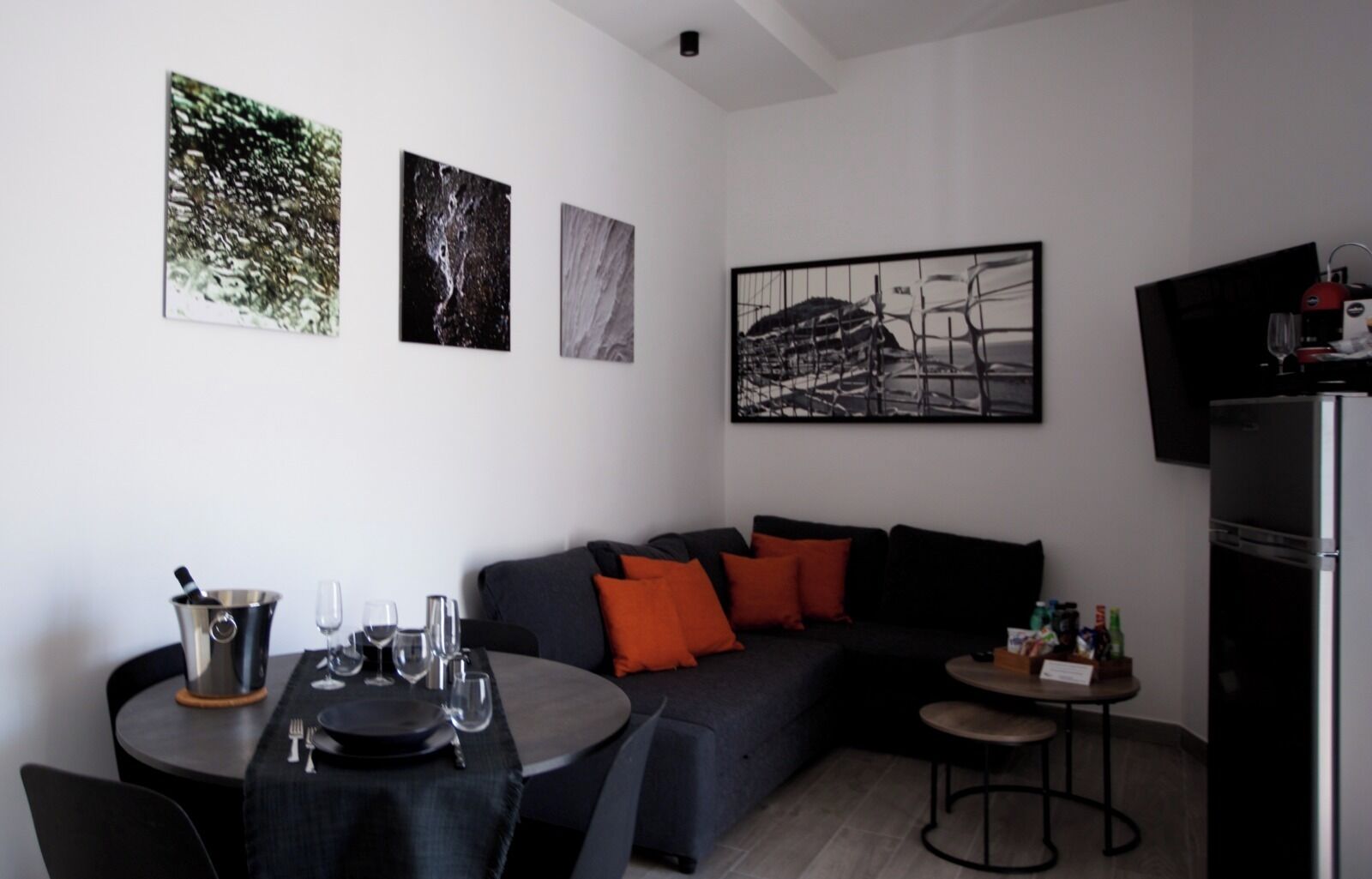 A modern living room with a dark grey sofa, orange cushions, a dining table with wine glasses, and a small bar area.