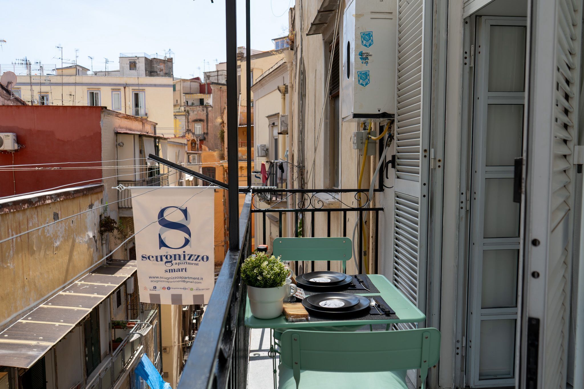 Ein schmaler Balkon mit einem kleinen Tisch, einem Stuhl und einem Blumentopf. Ein Schild mit der Aufschrift 'Scugnizzo apartment smart' ist sichtbar.