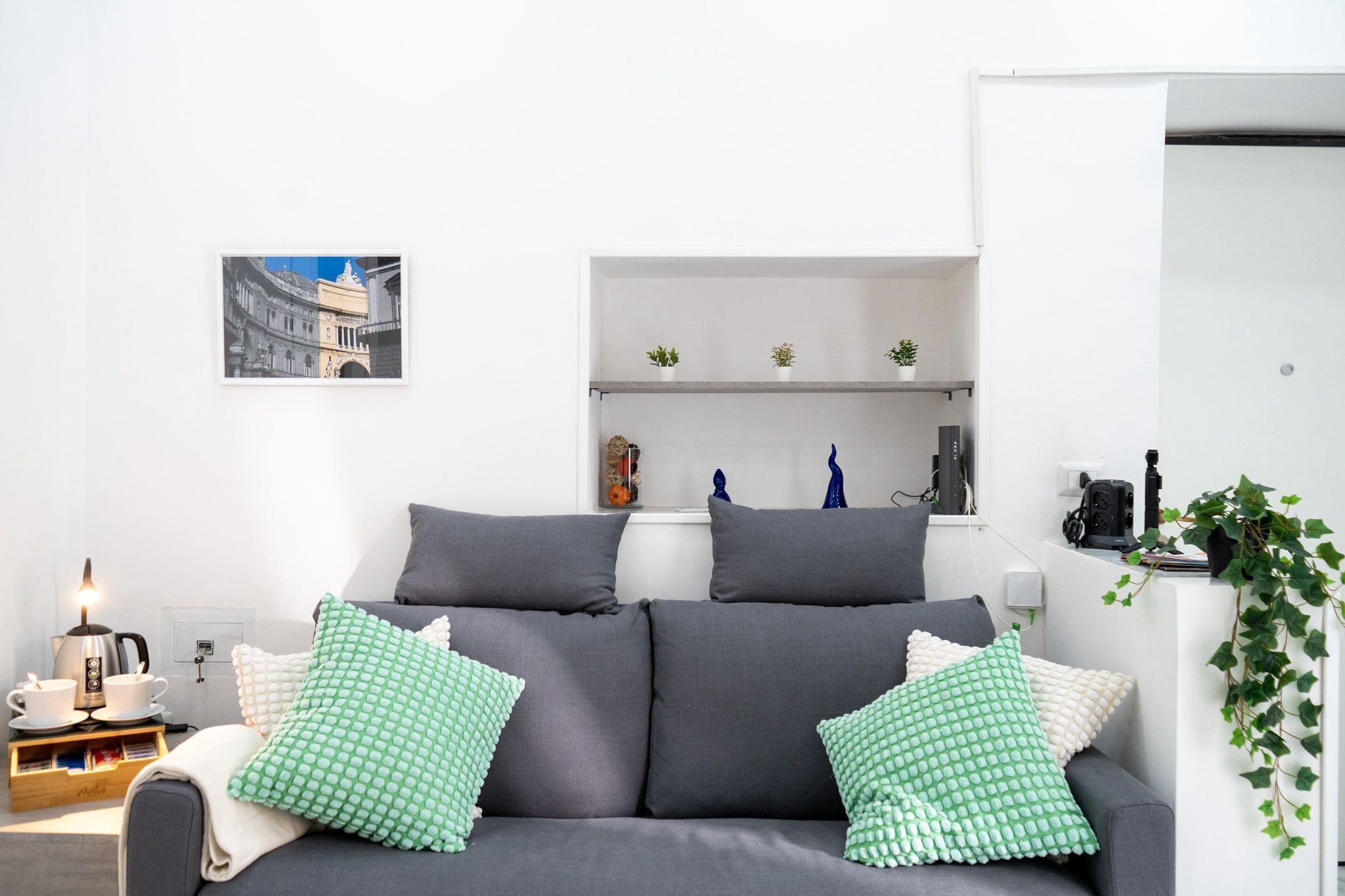 Un salon moderne avec un canapé gris, des coussins à motifs verts et blancs, une table basse avec une bouilloire, un tableau encadré au mur et une petite étagère avec des objets décoratifs.
