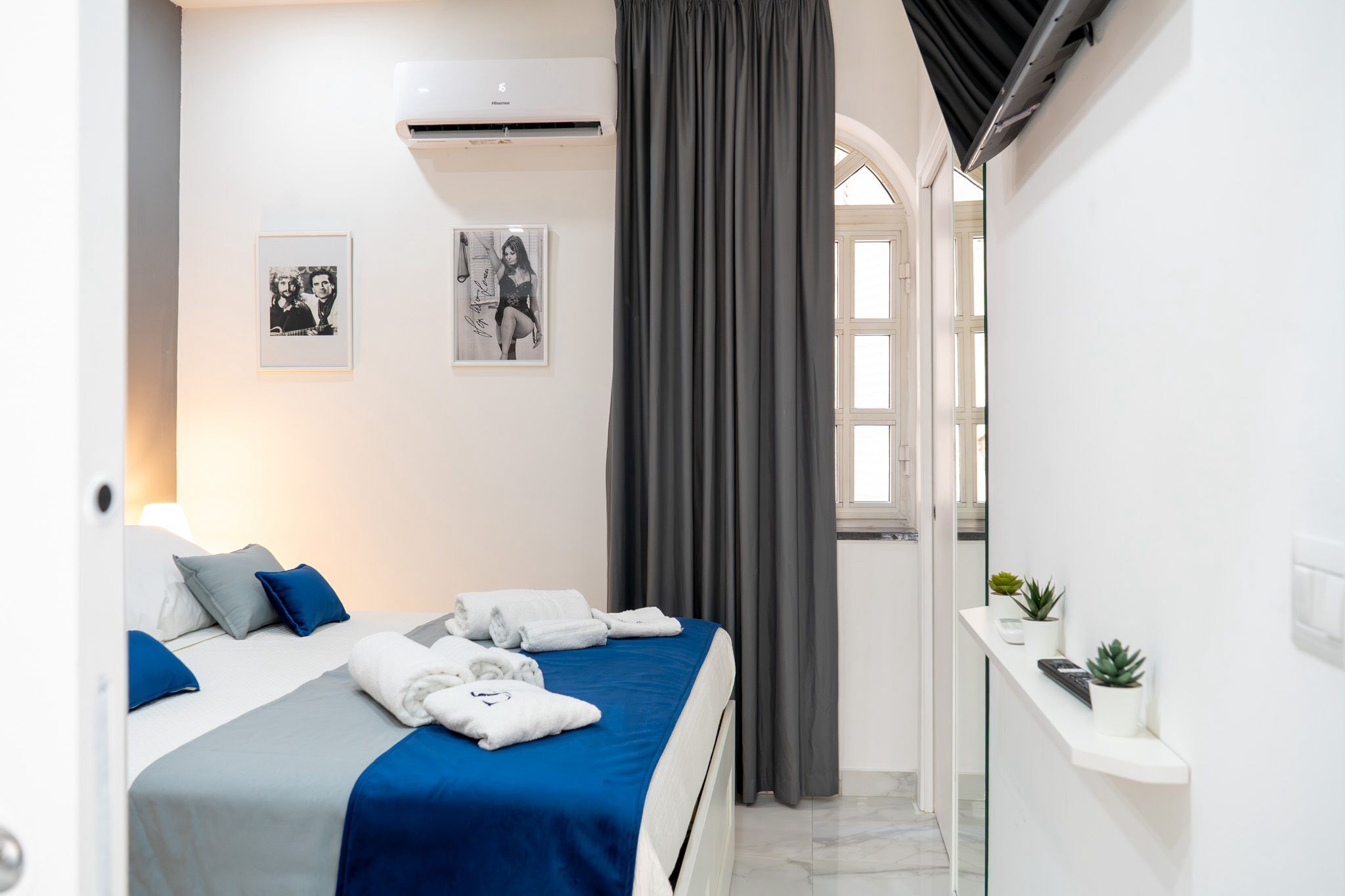 Una habitación de hotel con una cama, dos almohadas, una colcha azul, una cortina gris, un aire acondicionado instalado en la pared y dos cuadros enmarcados.