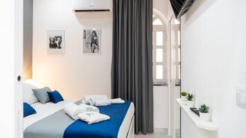 Una habitación de hotel con una cama, dos almohadas, una colcha azul, una cortina gris, un aire acondicionado instalado en la pared y dos cuadros enmarcados.