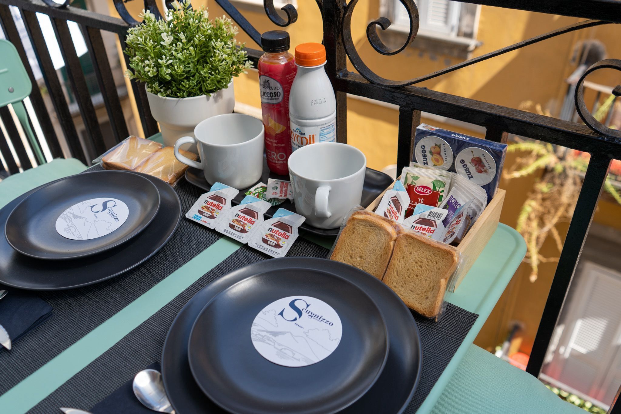 A table set with plates, cups, and food items, including bread and Nutella packets, on a balcony with a potted plant and bottles in the background.