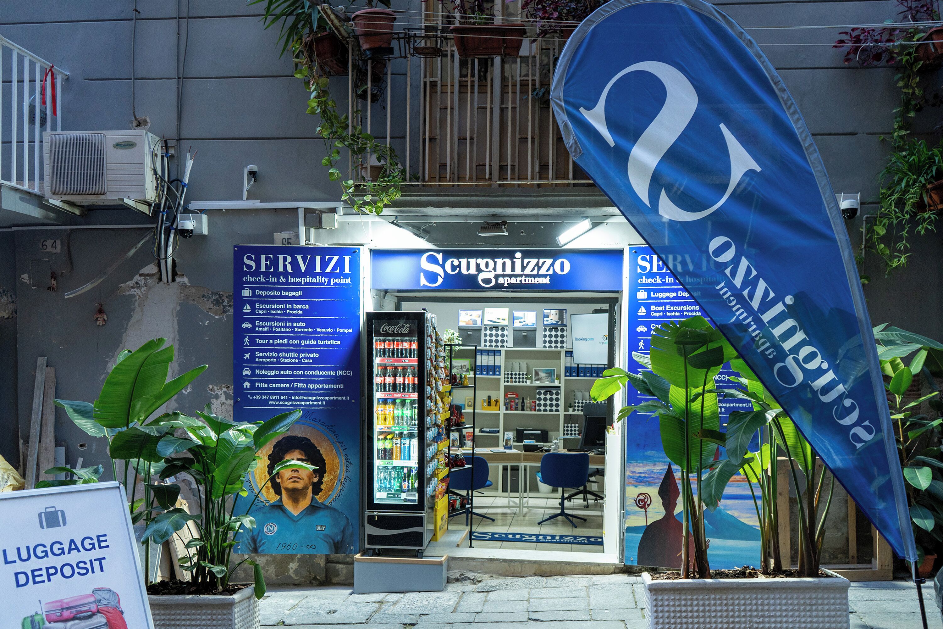 A hotel entrance with a Coca-Cola vending machine, a 'Luggage Deposit' sign, and a 'Servizi' sign indicating check-in and hospitality services.