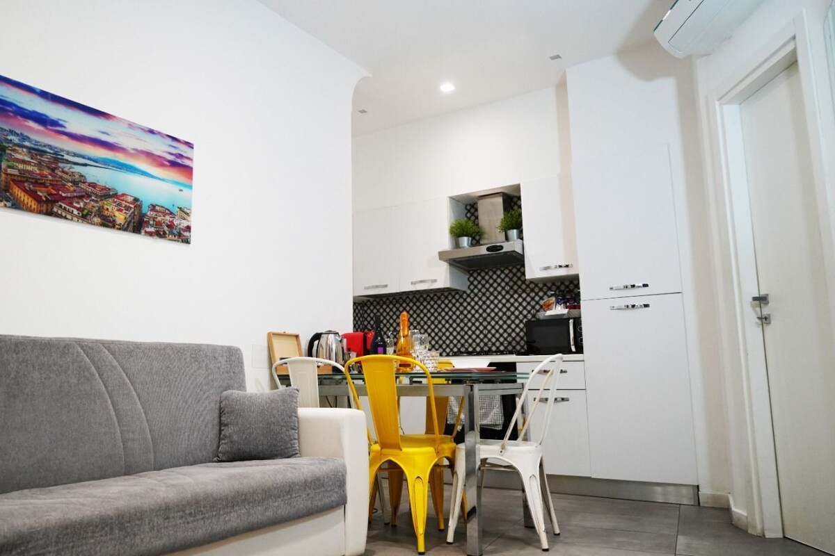 A living room with a grey sofa, a dining table with yellow chairs, and a kitchen area with white cabinets and a black backsplash.
