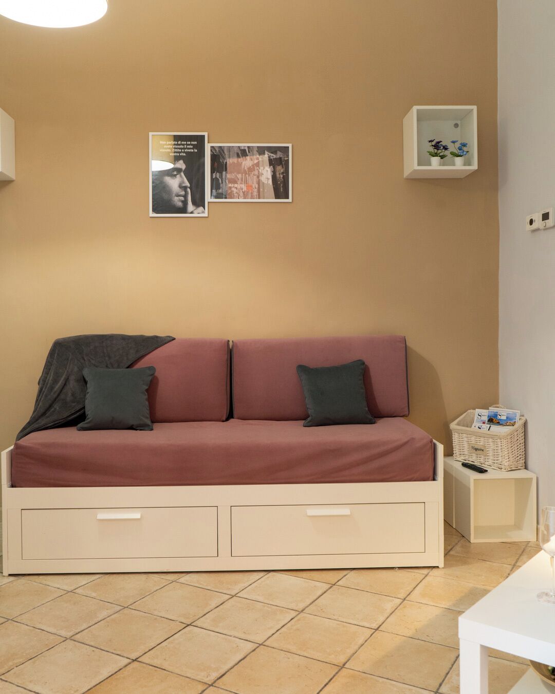 En moderne stue med en beige sofa, to puder, et lille hvidt sofabord, en kurv til magasiner og en fjernbetjening.
