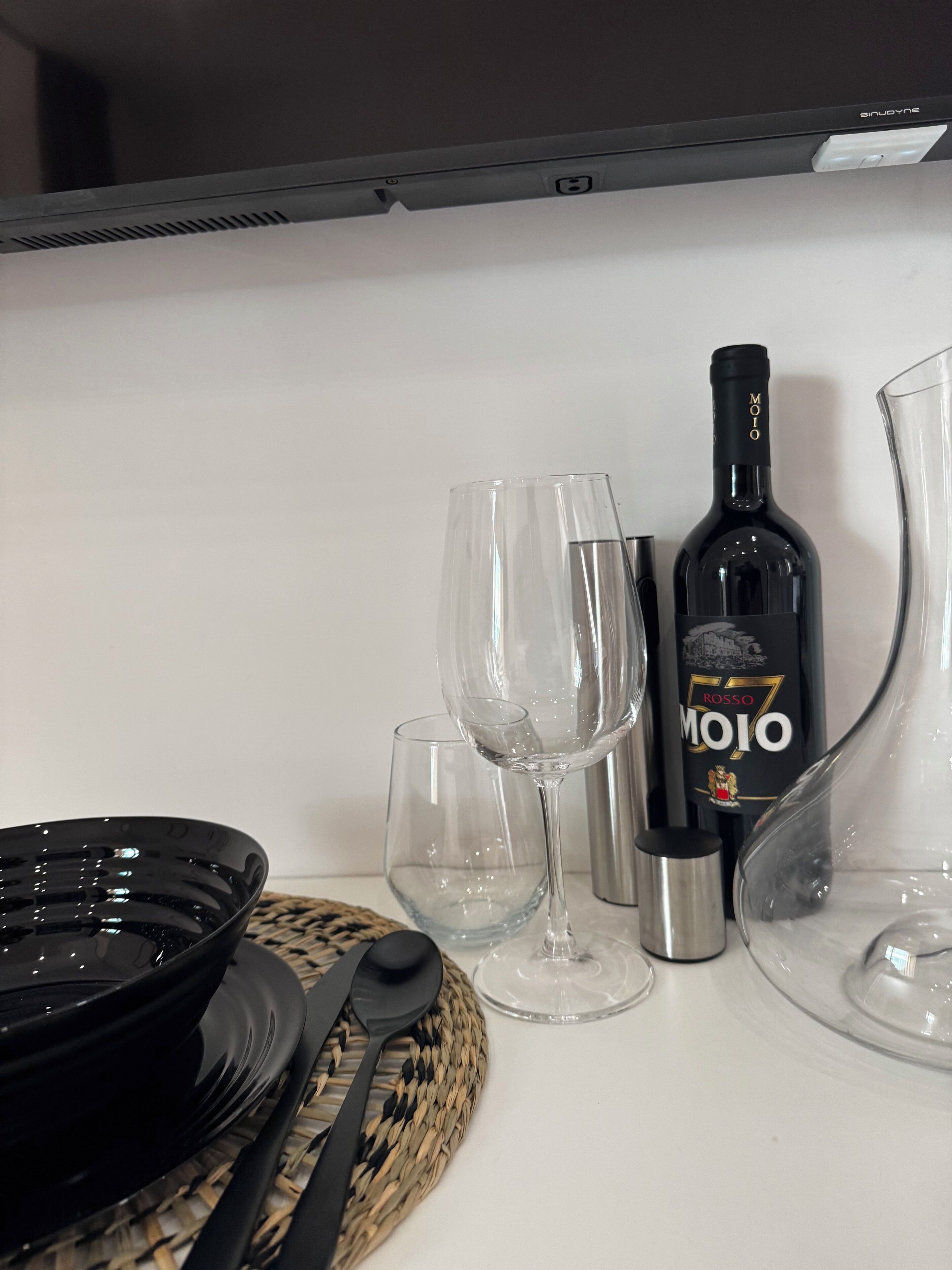 A dining setup with a black plate, clear wine glasses, a wine bottle, and a stainless steel tumbler on a white table.