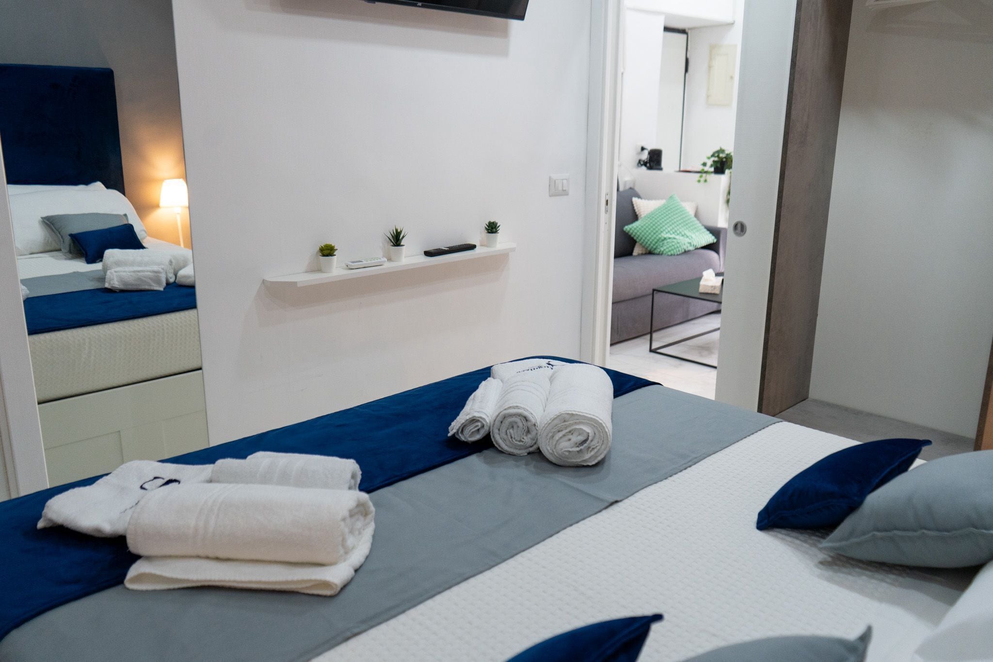 Een hotelkamer met een bed, een klein plankje met planten en uitzicht op een andere kamer.