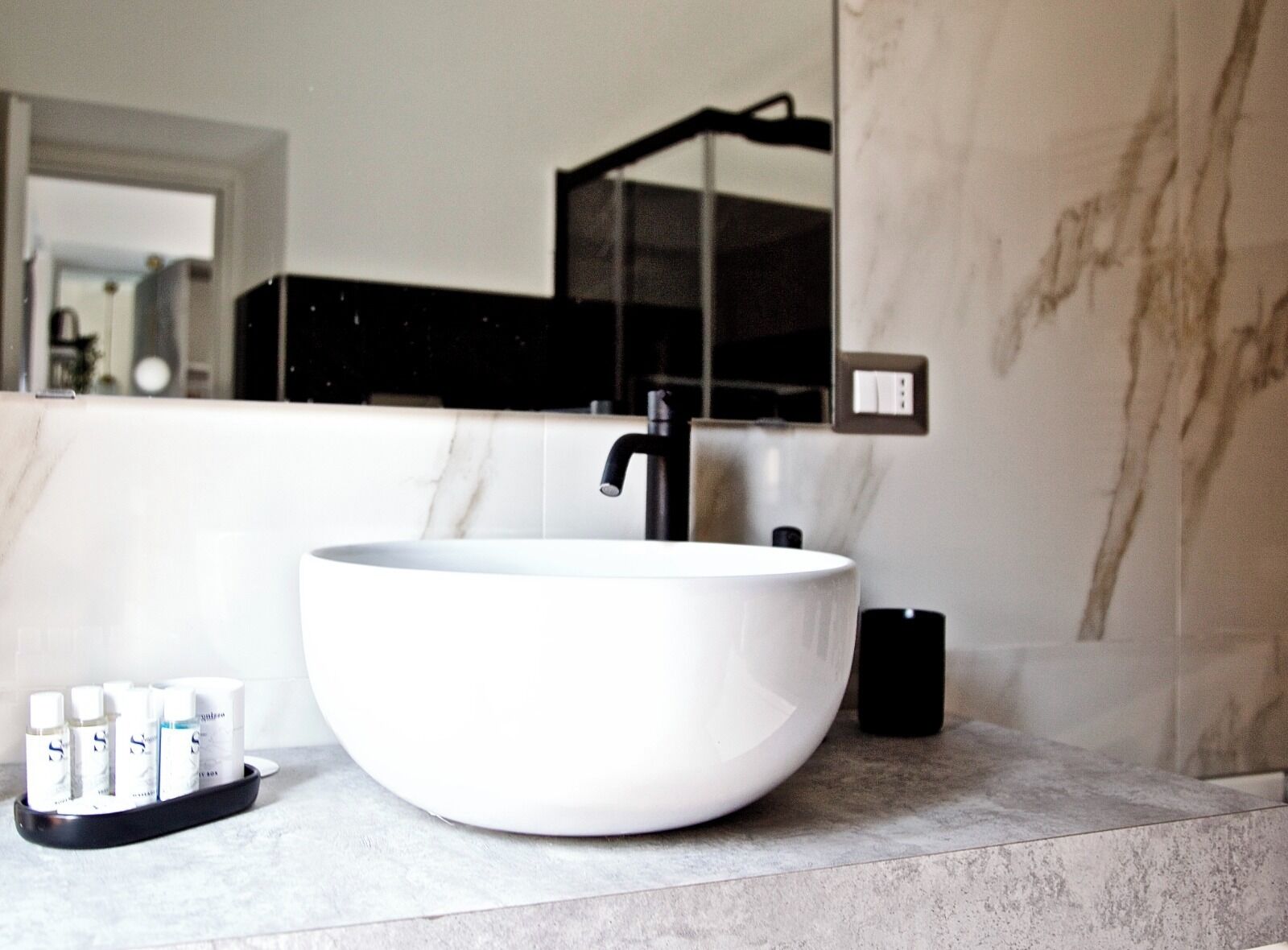 A modern bathroom with a large white vessel sink, a black faucet, and a mirror above it. There are toiletries on a tray next to the sink.