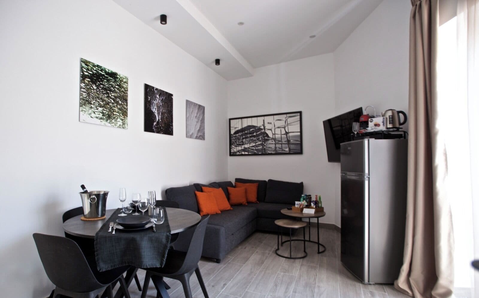 Ein modernes Wohnzimmer mit Essbereich, einer Couch mit orangefarbenen Kissen, einem kleinen runden Tisch mit Wasserkocher und einem Kühlschrank.