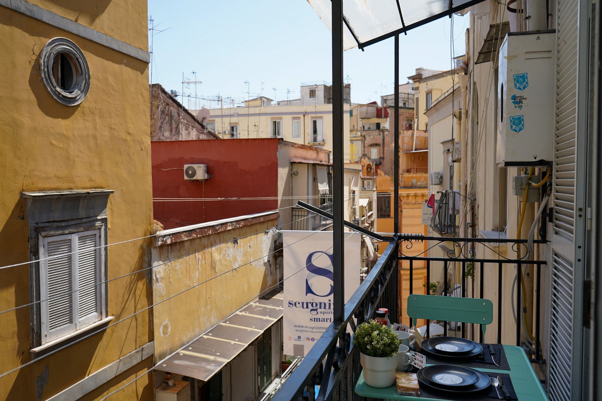 A narrow balcony with a metal railing, a small table with a potted plant, and a sign advertising 'Scugnizzi Smart'.