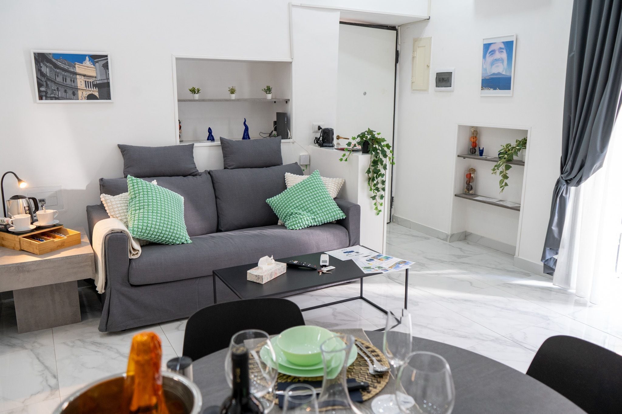 Un salón moderno con un sofá gris, cojines verdes y blancos, una mesa de centro y una zona de comedor con una mesa preparada para una comida.