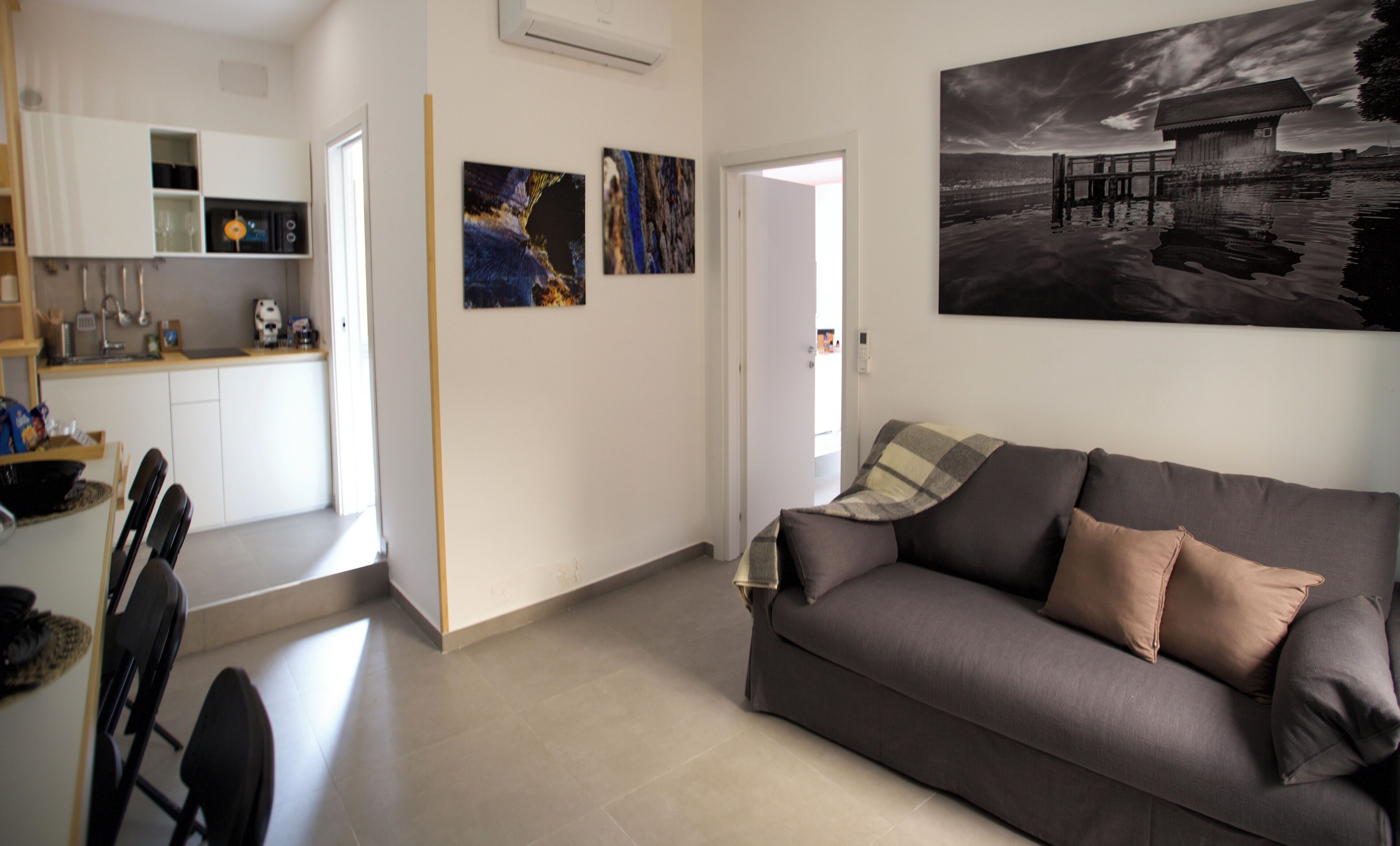 Een moderne woonkamer met een grijze bank, een eethoek met stoelen en een keuken op de achtergrond.