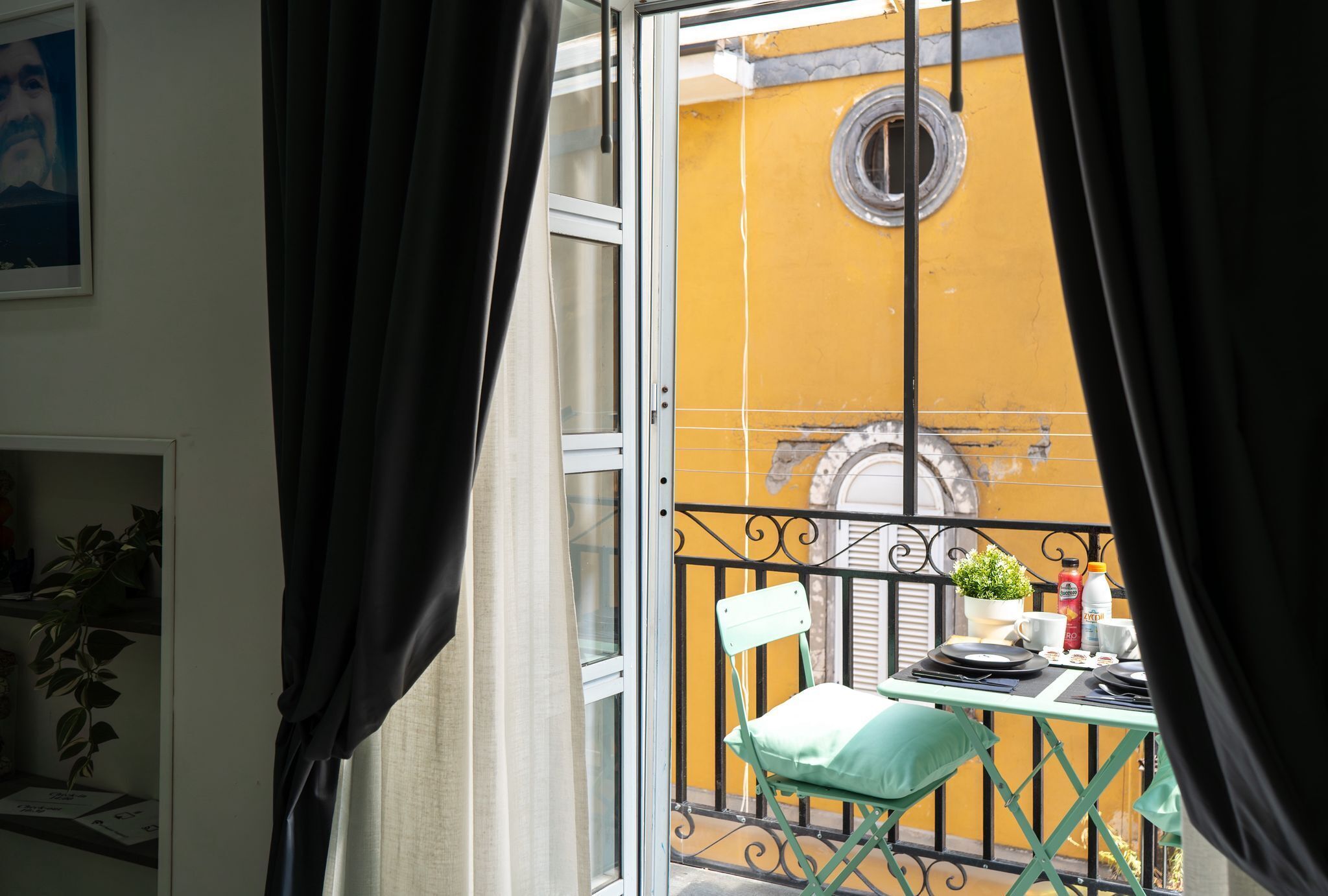 A balcony with a yellow wall, a metal railing, a table with a chair, and a window with curtains.