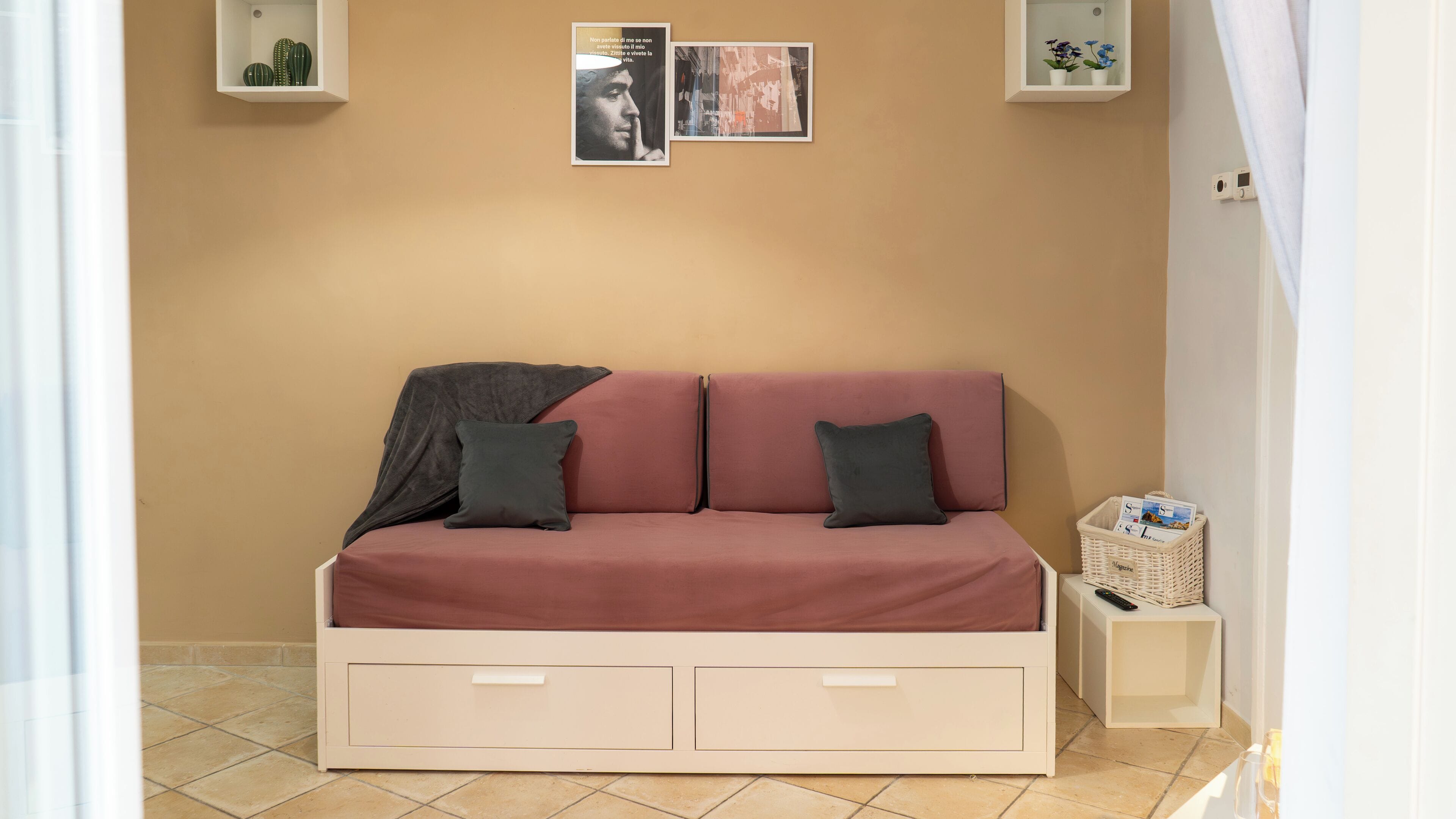 Une petite pièce accueillante, avec un canapé beige, deux coussins, une couverture et une petite table d’appoint blanche sur laquelle se trouvent un panier et une télécommande.