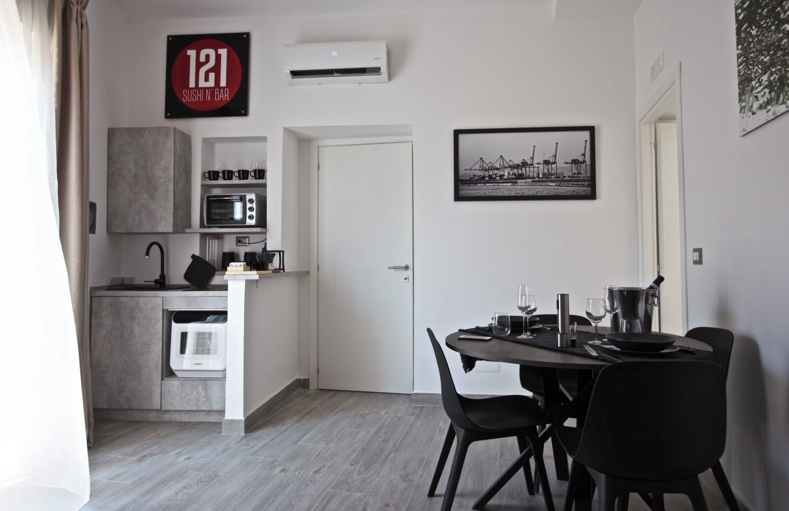 A compact kitchen with a microwave, oven, and a coffee machine. There is a dining area with a table set for two and a black chair. A framed picture hangs on the wall, and a sign above the door reads "121 SUSHI N' BAR".