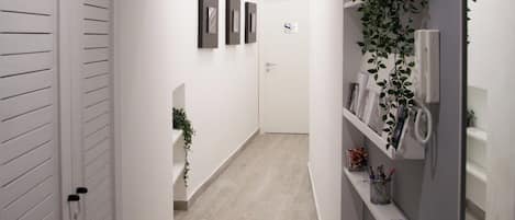 A narrow hallway with white walls, a light wooden floor, and a white door at the end. There are three framed pictures on the wall, a small plant, and a shelf with various items.