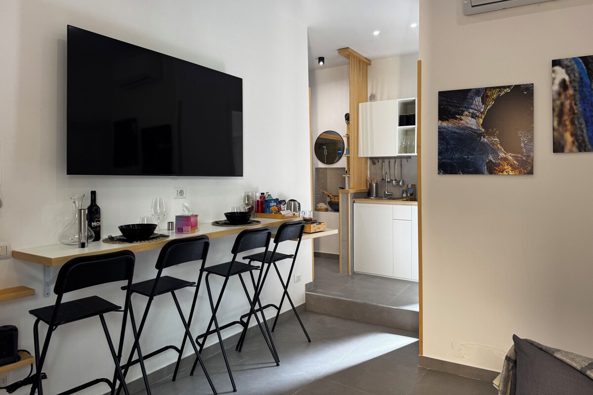 A modern kitchen with a flat-screen TV, a dining area with four chairs, and a small bar area.
