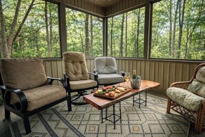 A covered porch with four chairs, a wooden table with a fruit platter, and a bottle of wine.