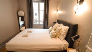 Ein ordentlich bezogenes Bett mit Kissen und einer Nachttischlampe in einem Zimmer mit Fenster und Wandleuchte.