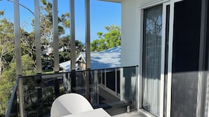 Un balcón con barandilla de cristal, una silla blanca y vistas a una casa con tejado azul.