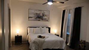 Een keurig opgemaakt bed met witte lakens, twee nachtkastjes met lampen, een plafondventilator en een groot abstract schilderij aan de muur.