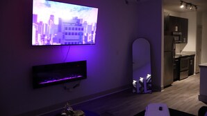 A modern living room with a flat-screen TV mounted on the wall, a small shelf below it, and a kitchen visible through an open doorway. - Modern Midtown 1BR VIP World Cup Experience (Atlanta)