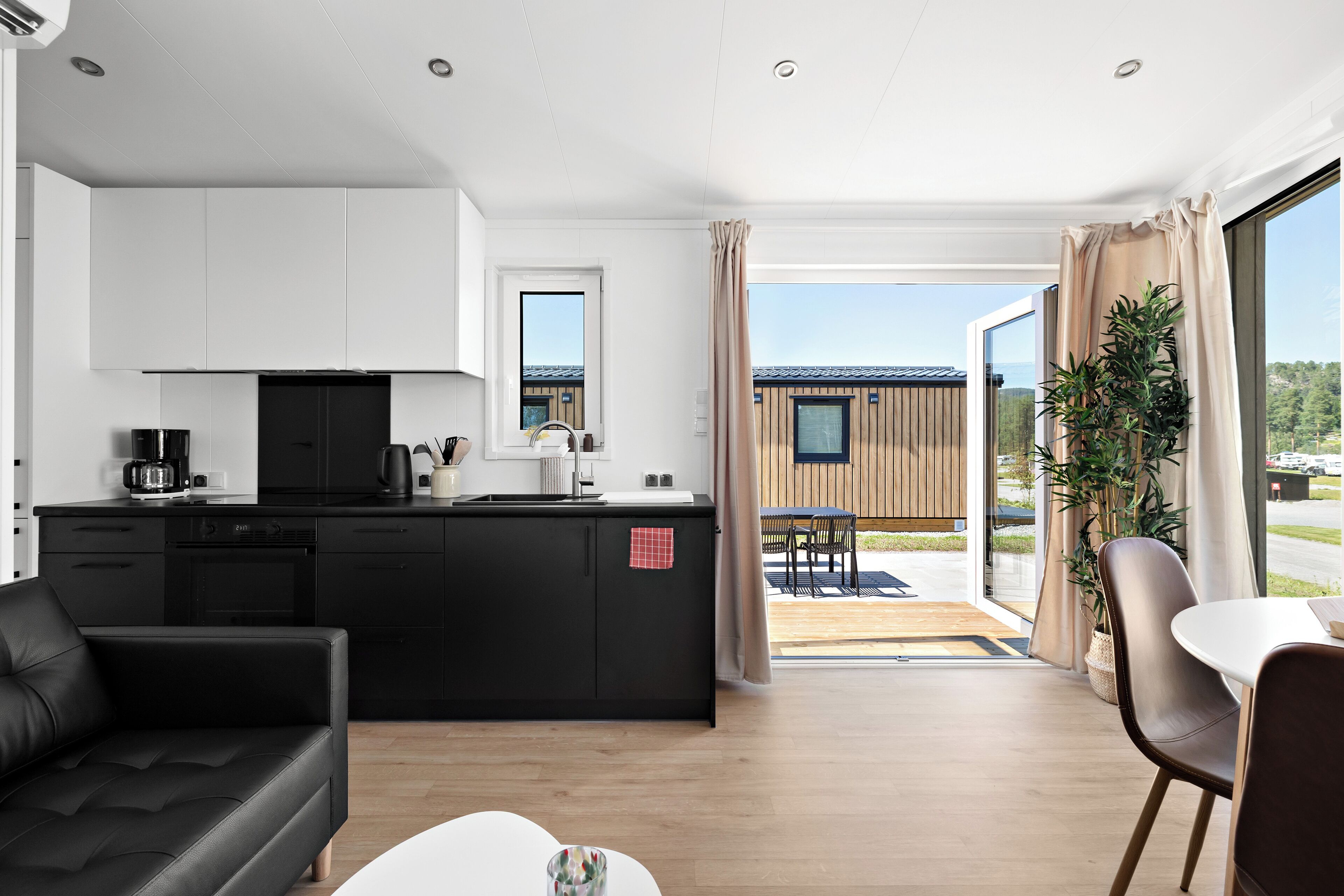 Eine moderne Küche mit weißen Schränken, einer schwarzen Arbeitsplatte und einem schwarzen Sofa. Es gibt eine Essecke mit Tisch und Stühlen sowie einen Blick ins Freie.