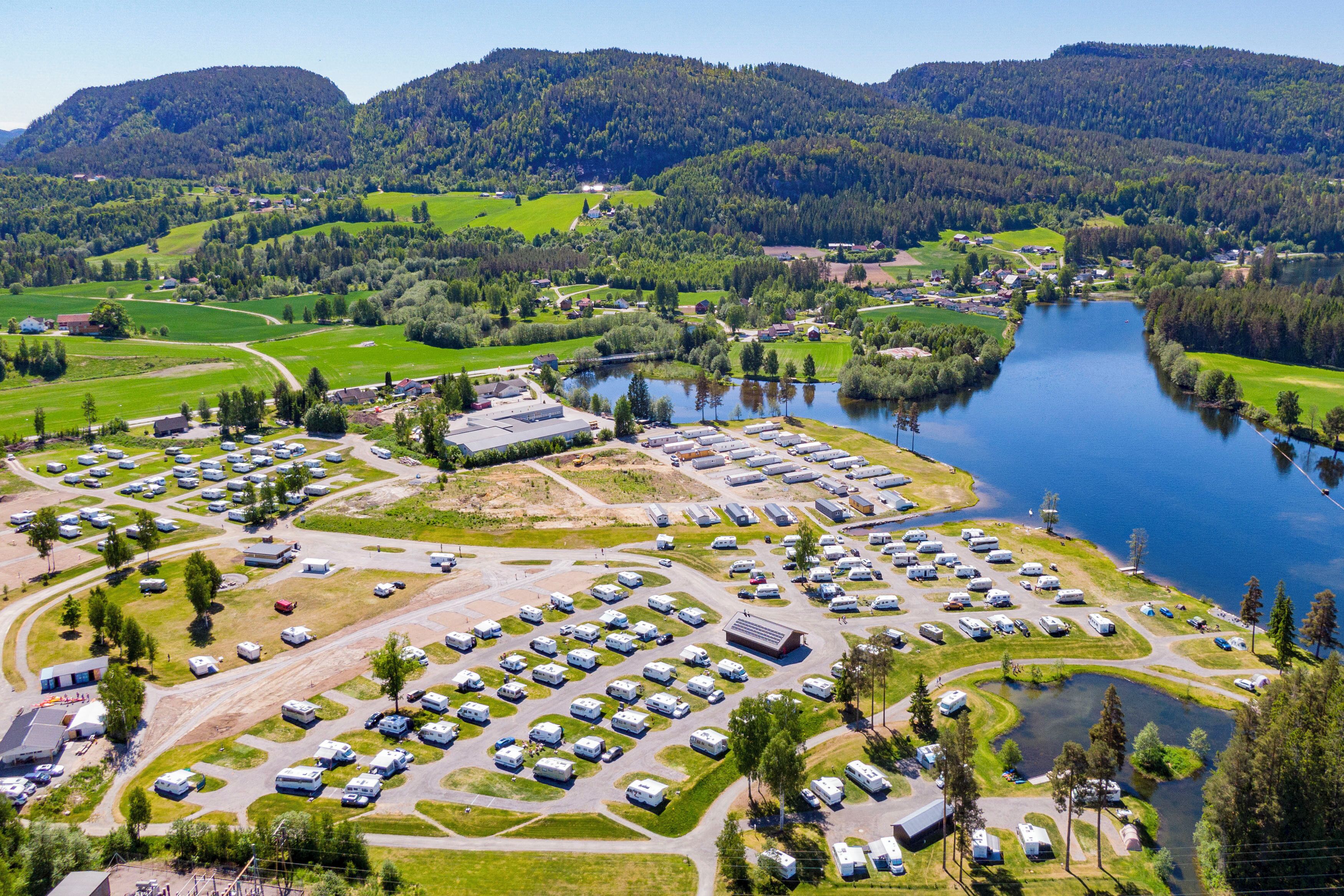 An aerial view of a campground with RVs parked in organized rows, a lake, and surrounding green hills.