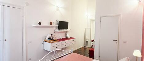 Un dormitorio con techo de ladrillo, paredes blancas, cama con ropa de cama rosa y blanca, cómoda blanca con televisor y una silla roja.