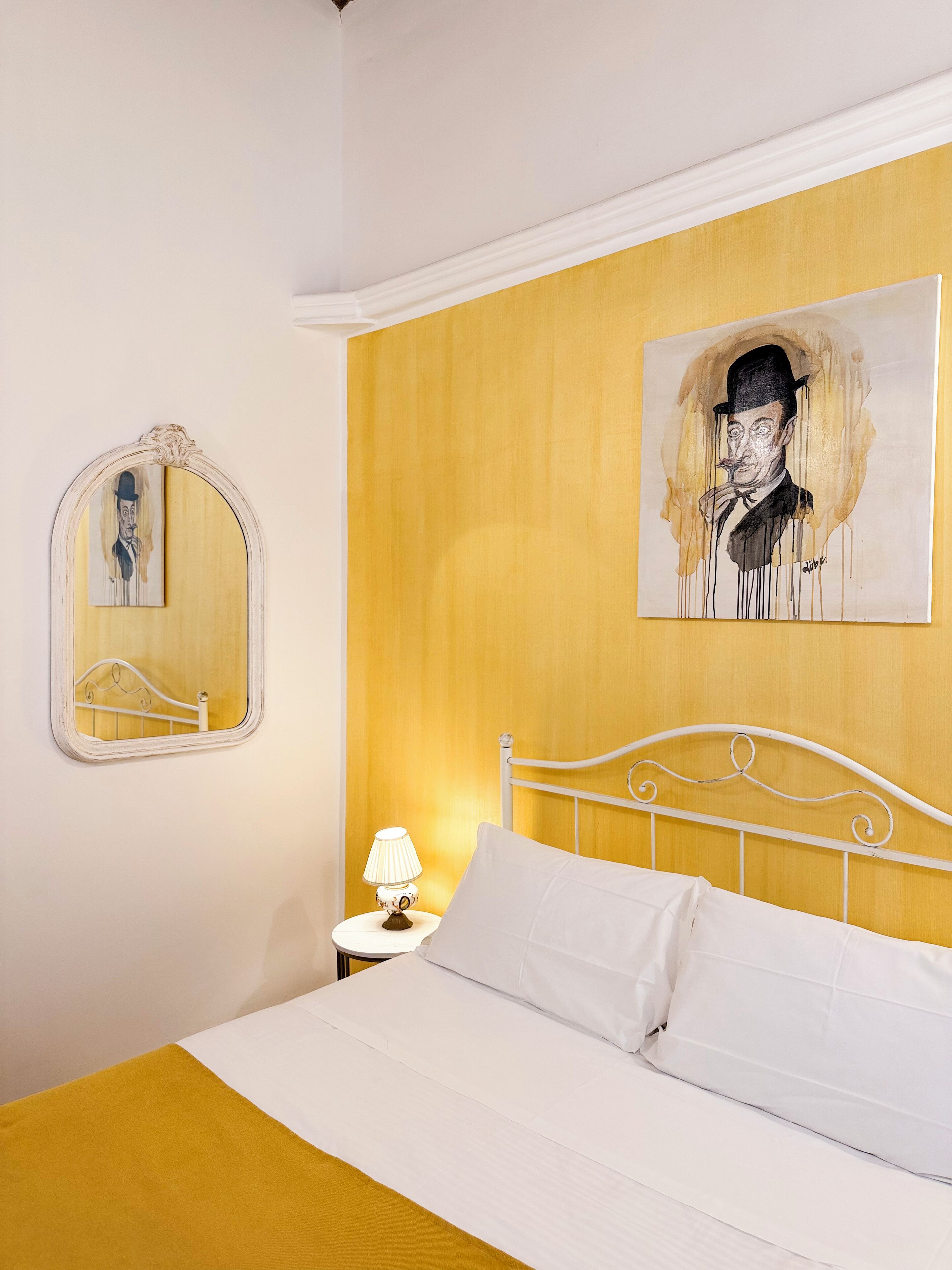 A bedroom with a bed, a bedside table with a lamp, a mirror, and a painting on the wall.