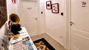 Un couloir avec une horloge, des tableaux encadrés, une porte portant l’inscription « CHAMBRE Estinotte » et une table sur laquelle se trouvent une carte et divers objets.