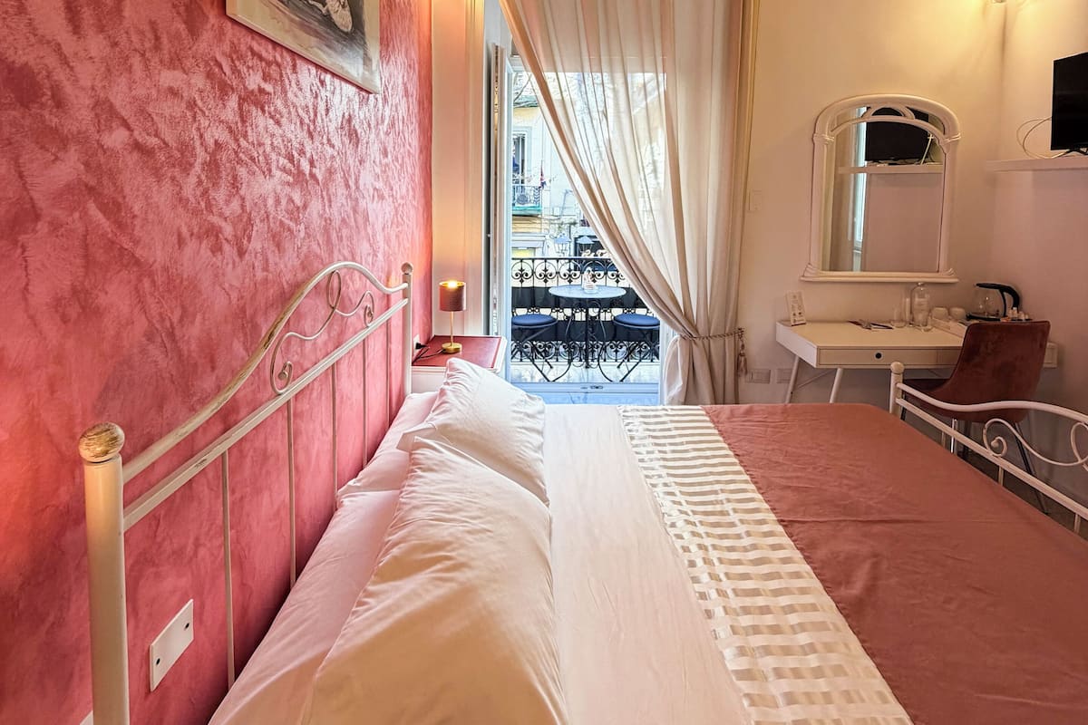 A bedroom with a bed, a desk, a chair, a mirror, and a view of the outside balcony.