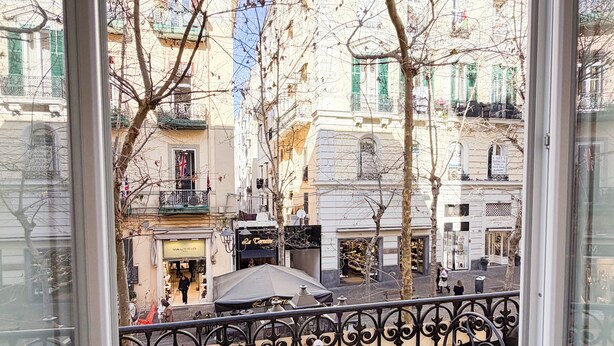 Un balcon avec une table ronde et deux chaises, donnant sur une rue bordée d’immeubles et d’arbres sans feuilles.