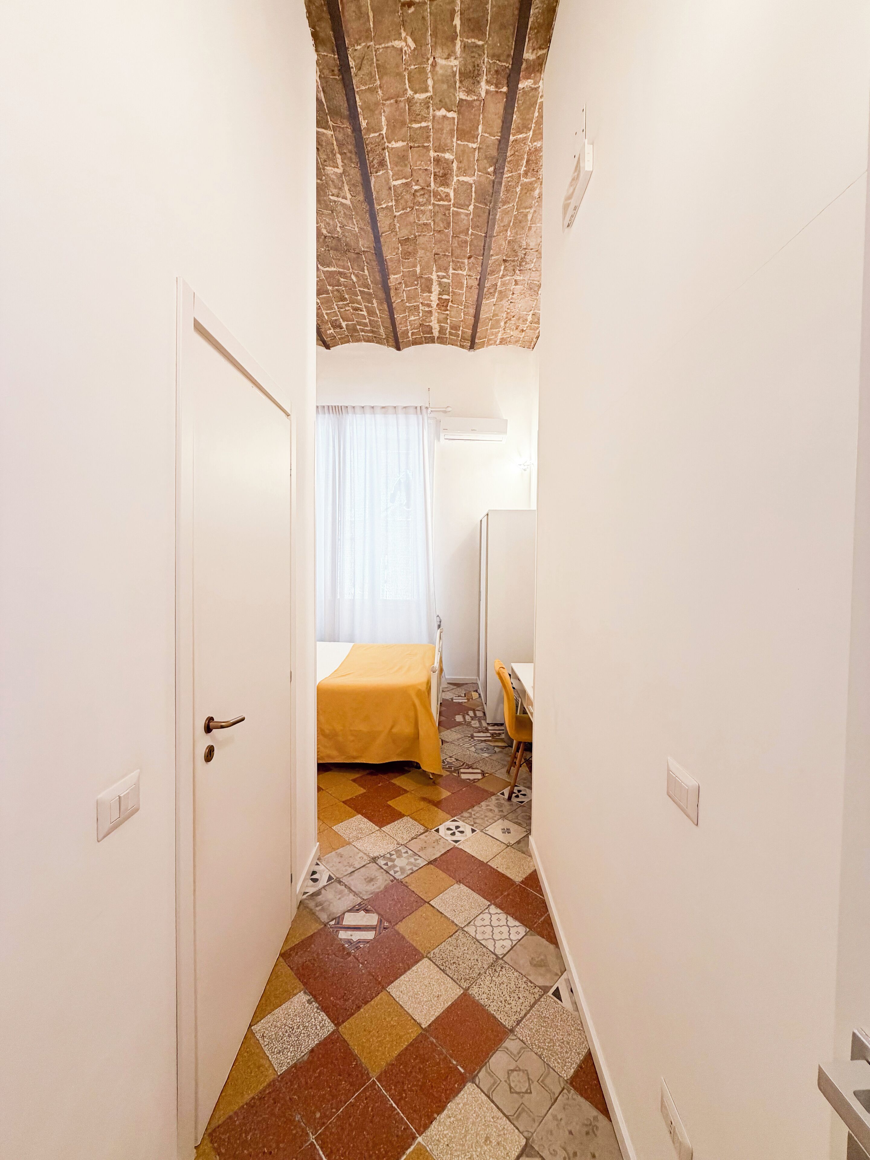 Un couloir étroit mène à une chambre qui contient un lit, une chaise et un bureau. La pièce est dotée d’un sol carrelé et d’un plafond orné d’un motif en briques.