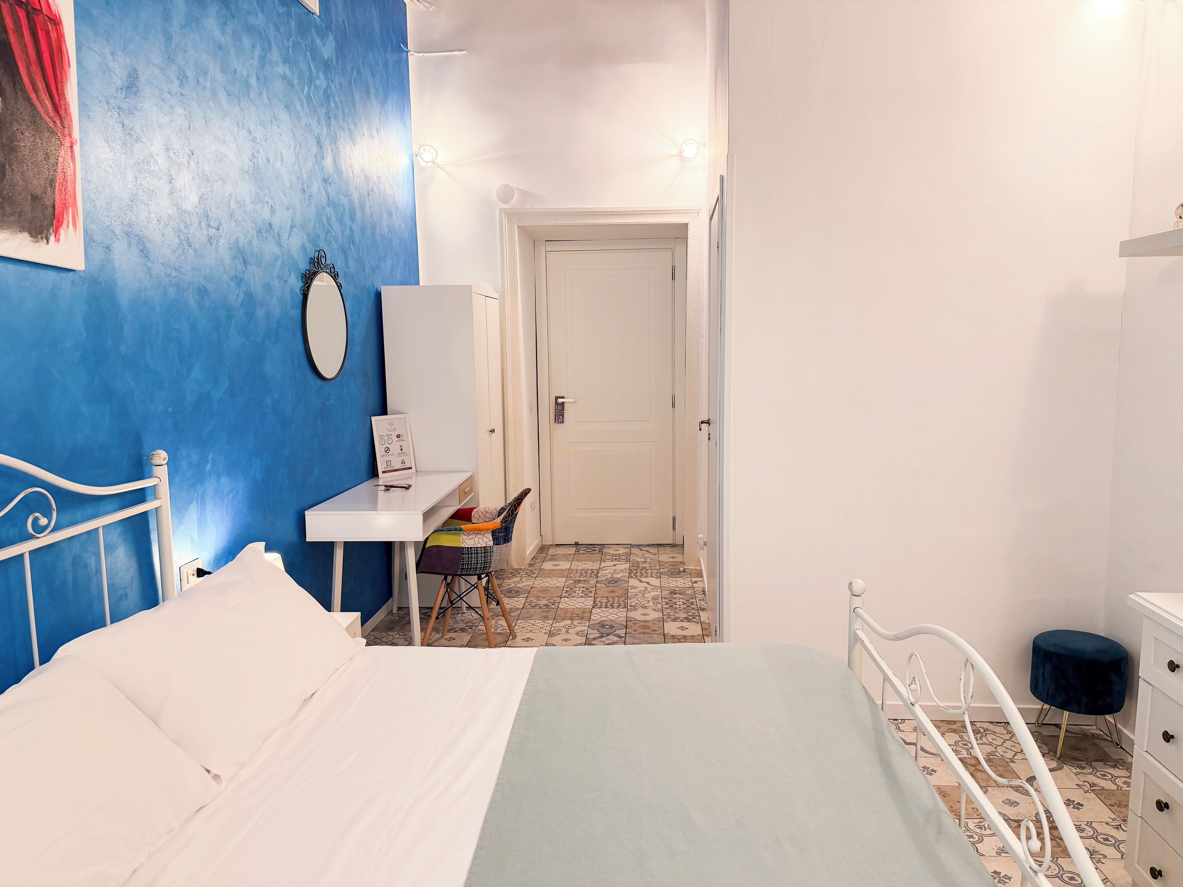 A bedroom with a bed, a desk, a chair, a dresser, and a door leading to another room.
