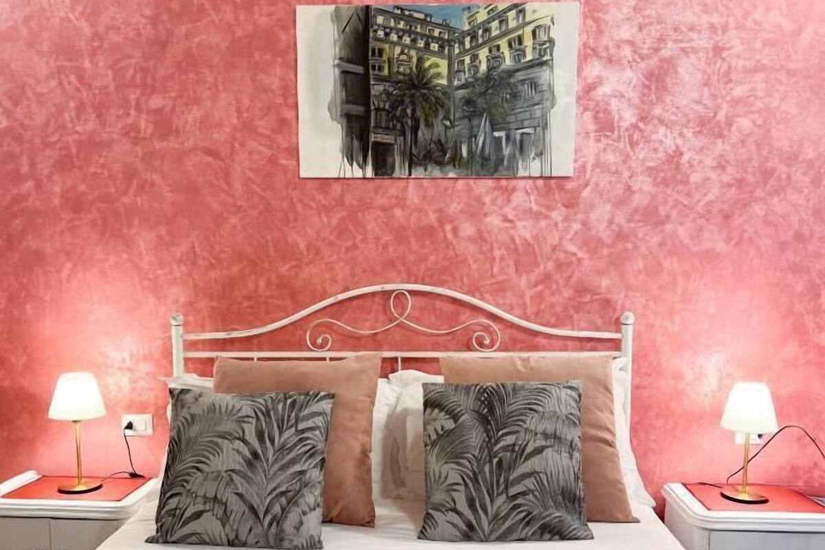 A bedroom with a red textured wall, a white bed with two pillows, and a painting of a cityscape above the bed.