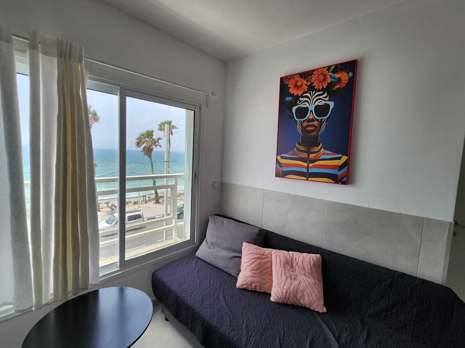 A room with a view of the ocean, a sofa with pillows, a painting on the wall, and a small table.