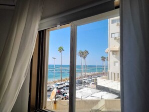 Blick aus einem Fenster auf einen Strand mit Palmen, einen Parkplatz und ein Gebäude mit Klimaanlagen.