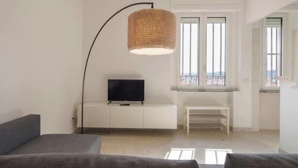 A modern living room with a flat-screen TV on a white cabinet, a large woven floor lamp, and a bench by the window. - Apartment with Panoramic Sea View in the Heart of Olbia (Olbia)