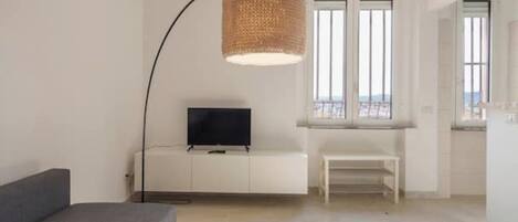 A modern living room with a flat-screen TV on a white cabinet, a large woven floor lamp, and a bench by the window.