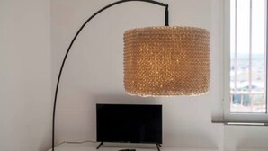 A modern room with a television on a white cabinet, a black floor lamp with a woven shade, and a window with a view of buildings. - Apartment with Panoramic Sea View in the Heart of Olbia (Olbia)
