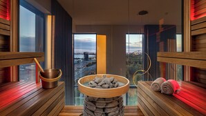 Una sauna con una panca in legno, una ciotola piena di pietre e vista sulla città attraverso ampie finestre.