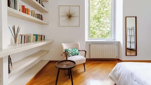 Ein Zimmer mit großem Fenster, einer Bücherwand, einem Sessel, einem runden Tisch und einem Bett.