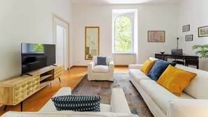 Ein Wohnzimmer mit Fernseher, einem weißen Sofa mit gelben Kissen, einem weißen Sessel und einem großen Fenster.