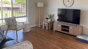 Een ruime woonkamer met een grote televisie, houten vloeren, een plafondventilator en uitzicht door grote ramen.