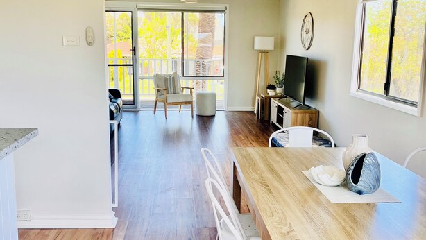 Een ruime eethoek met een houten tafel en witte stoelen, een televisie en uitzicht op de omgeving door grote ramen.