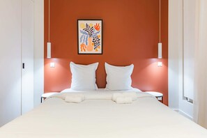 Ein Hotelzimmer mit einem großen Bett, weißer Bettwäsche, zwei weißen Kissen, einem gerahmten Kunstwerk an der Wand und Nachttischlampen.