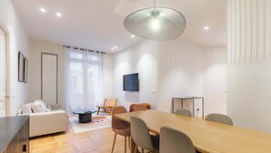 Ein modernes Wohnzimmer mit einer Couch, einem Esstisch mit Stühlen, einem an der Wand befestigten Fernseher und einem großen Fenster mit Vorhängen.
